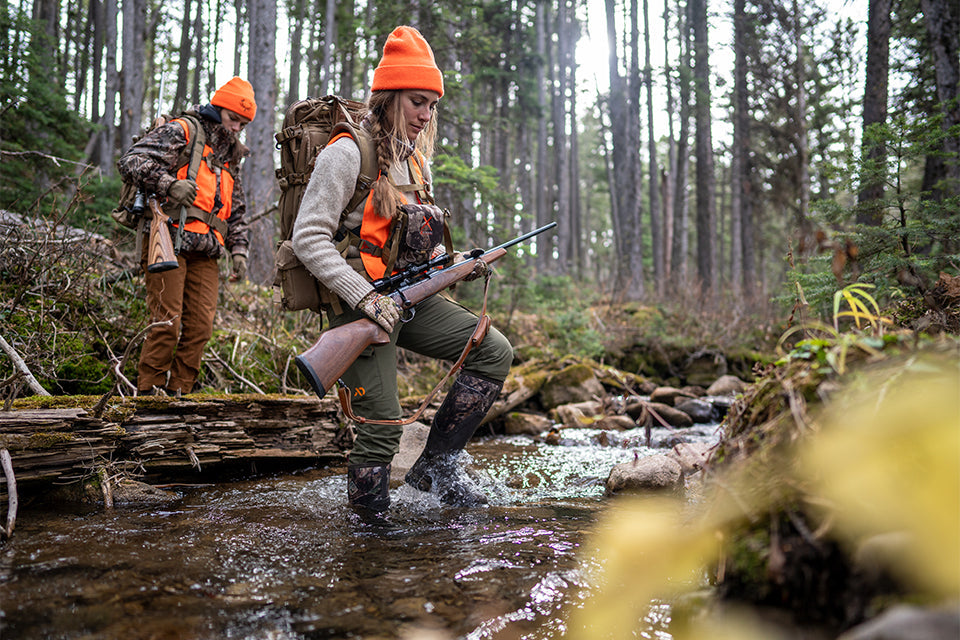 Woman crossing a creek, carrying rifle wearing hi vis hat and vest