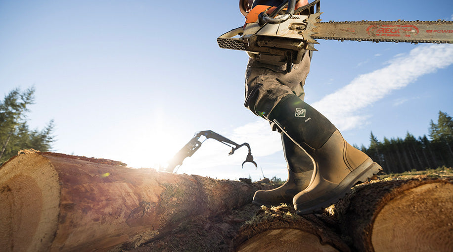 Man walking on cut tree log holding a chainsaw wearing Muck Chore Work Boots