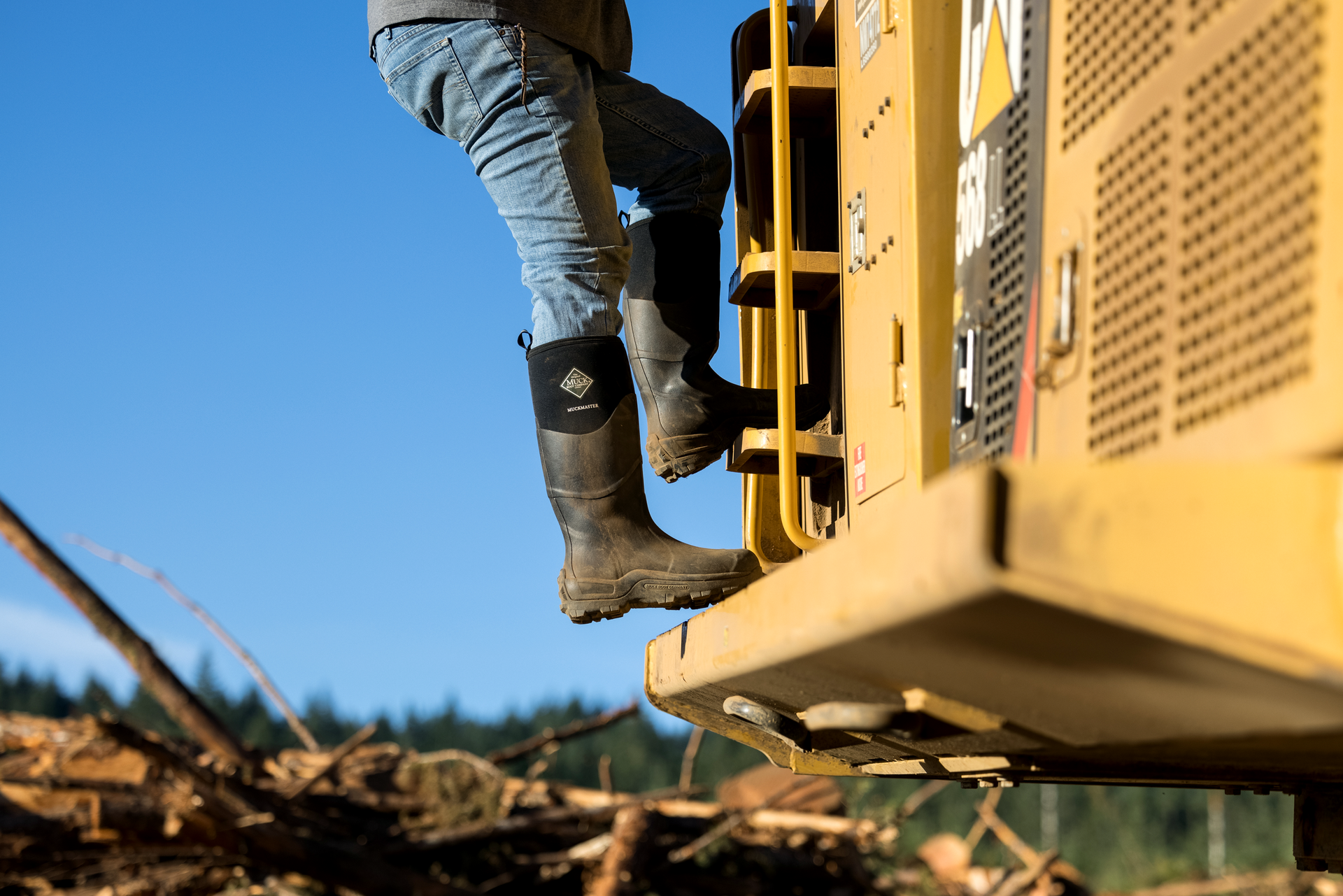 Person wearing rubber muck boots climbing onto a yellow piece of construction equipment with a clear blue sky in the background.