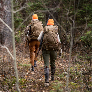 Two hunters walking through a forest with backpacks and orange hats in muck boots