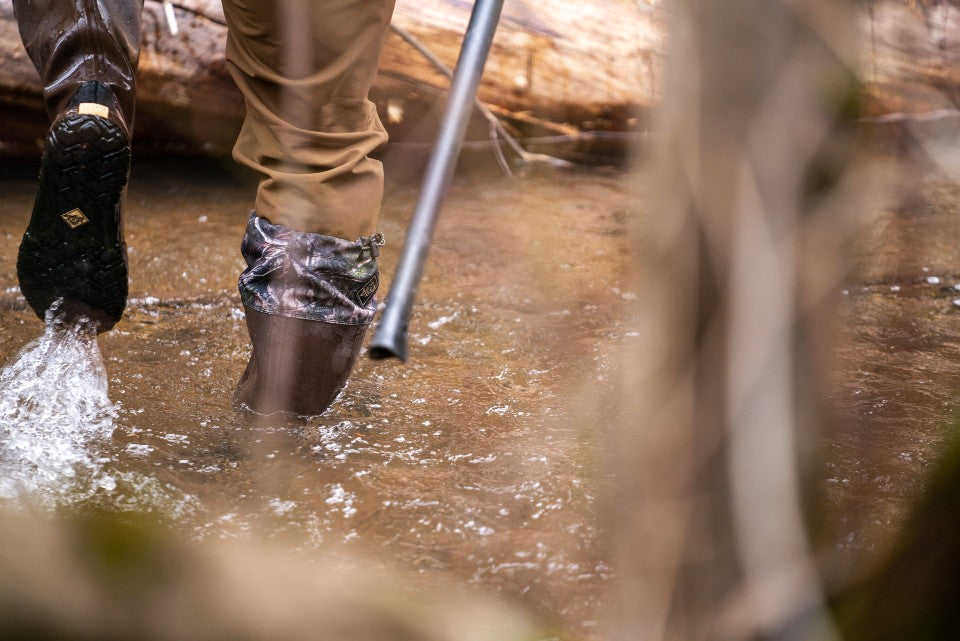 Person walking through a stream wearing knee high rubber muck boots.