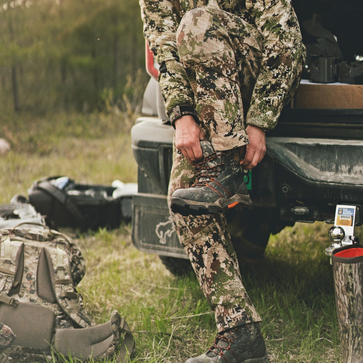 Person in camouflage gear preparing to hunt, standing next to a vehicle while pulling on muck boots