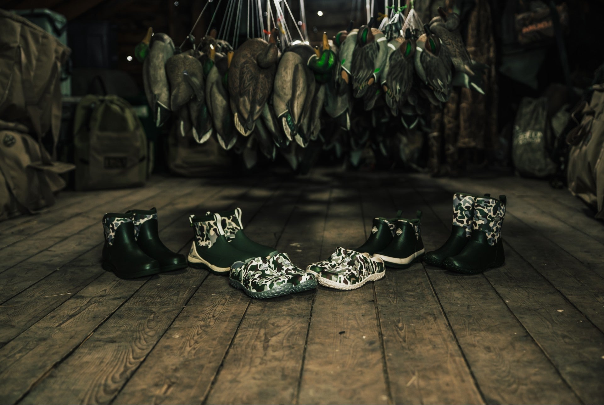 Muck Duck Camo printed footwear on a wooden floor with hanging decoys in the above