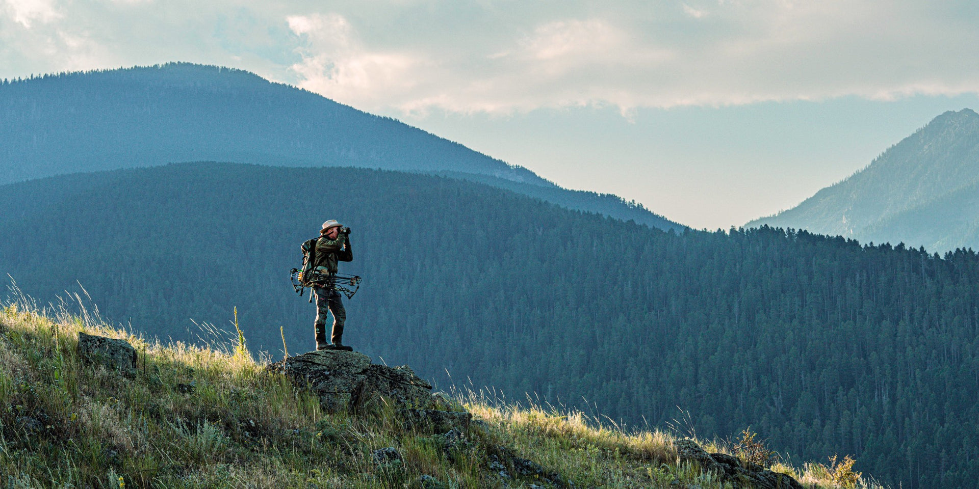 Jody Marler standing on a rocky outcrop with mountains in the background while elk hunting