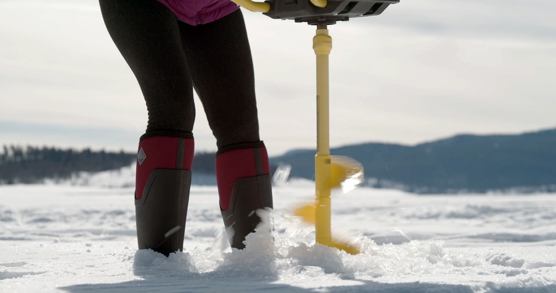 Person in winter muck boots using an ice auger to drill a hole in a frozen lake.