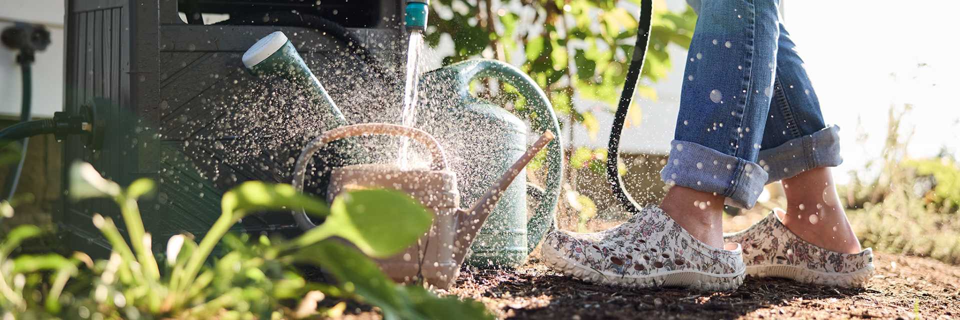 Person watering plants with a hose and watering can in a garden setting wearing Muckster Lite Women's Garden Clogs