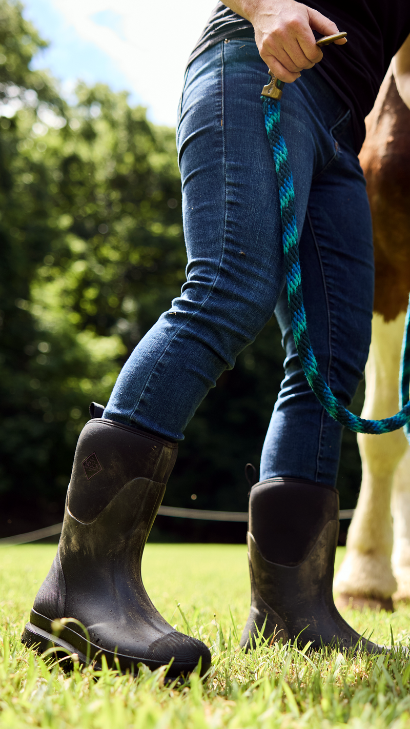 Person wearing blue jeans and black chore boots holding a horse's reins in a grassy area with trees in the background