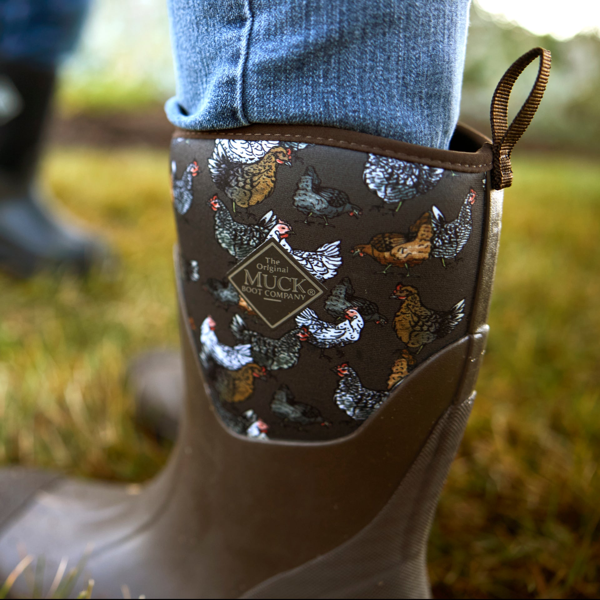 Close-up of a Women's Chore boot with a chicken pattern and Muck boot Company logo, worn outdoors.
