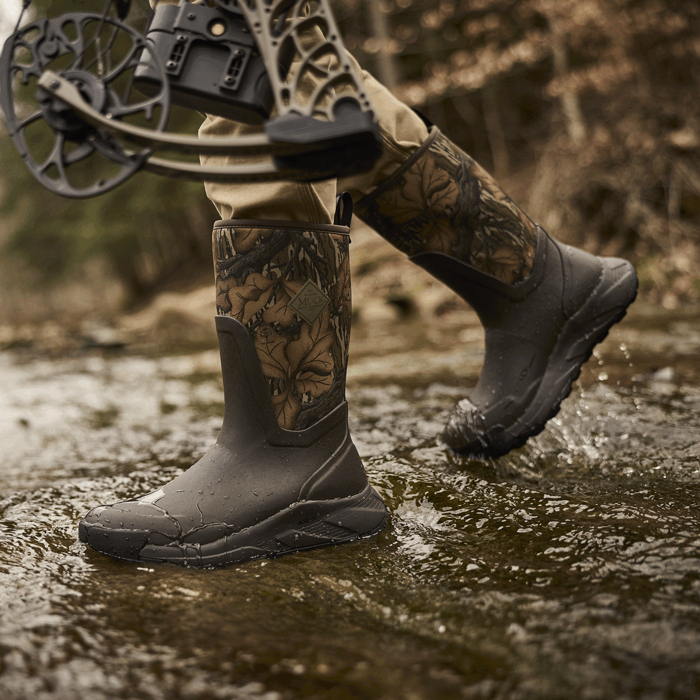 Person wearing Outmax 12" Camo Hunting boots in Mossy Oak Fall Foliage walking through water with a bow and arrow.