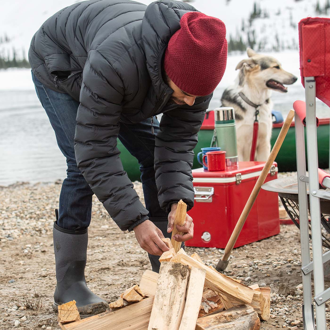 Person chopping wood by a lake with a dog and red cooler in the background