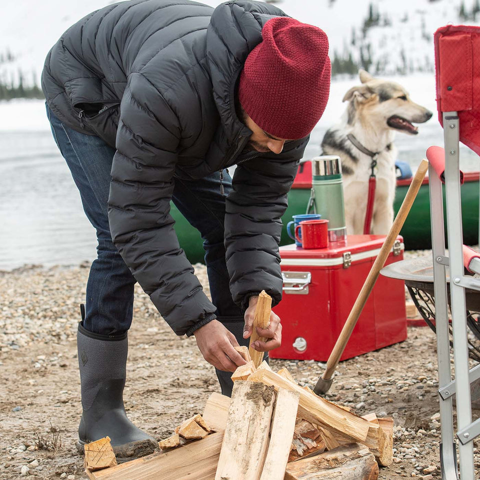 Person chopping wood by a lake with a dog and red cooler in the background