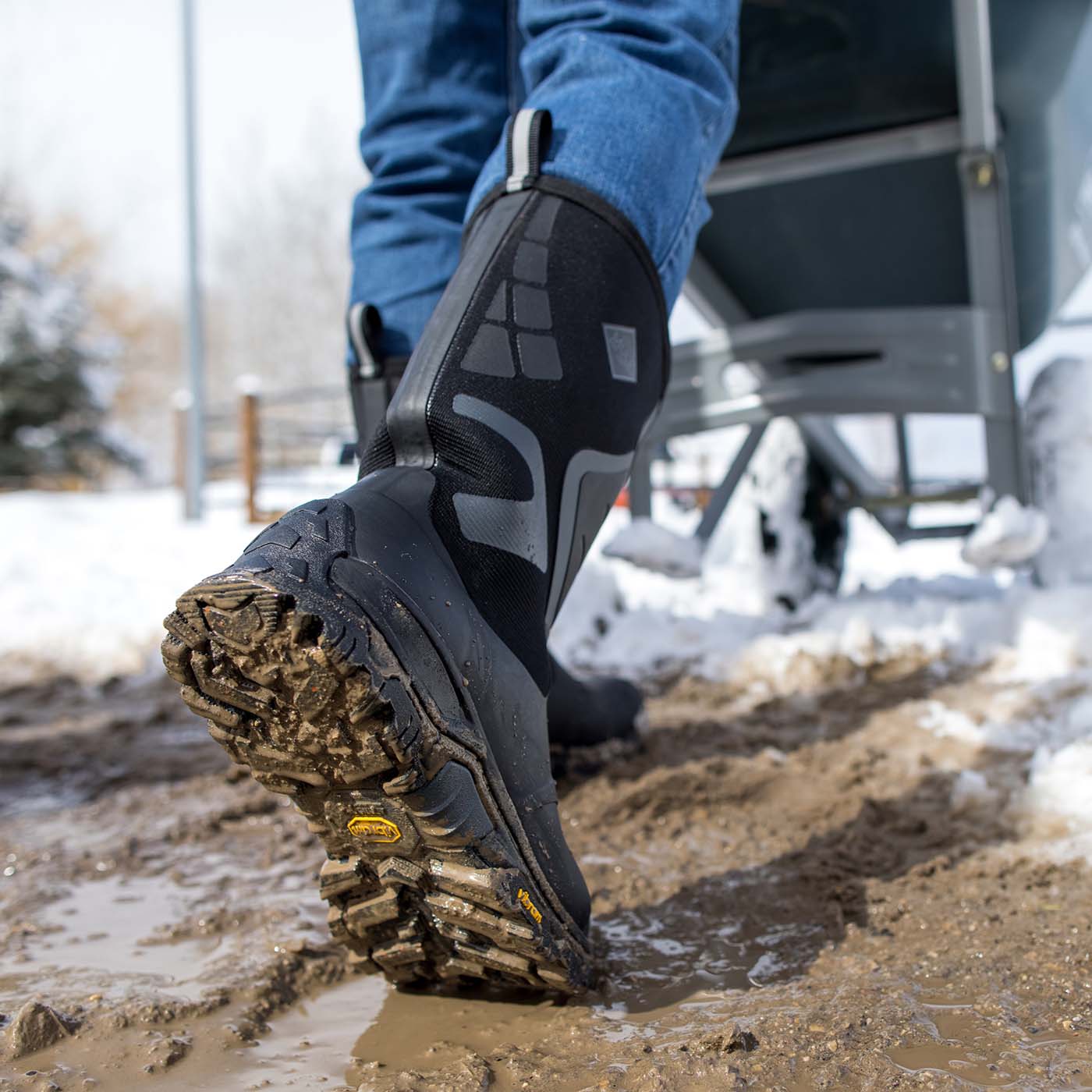 Person wearing black and gray rubber boots with a rugged outsole on a muddy path.