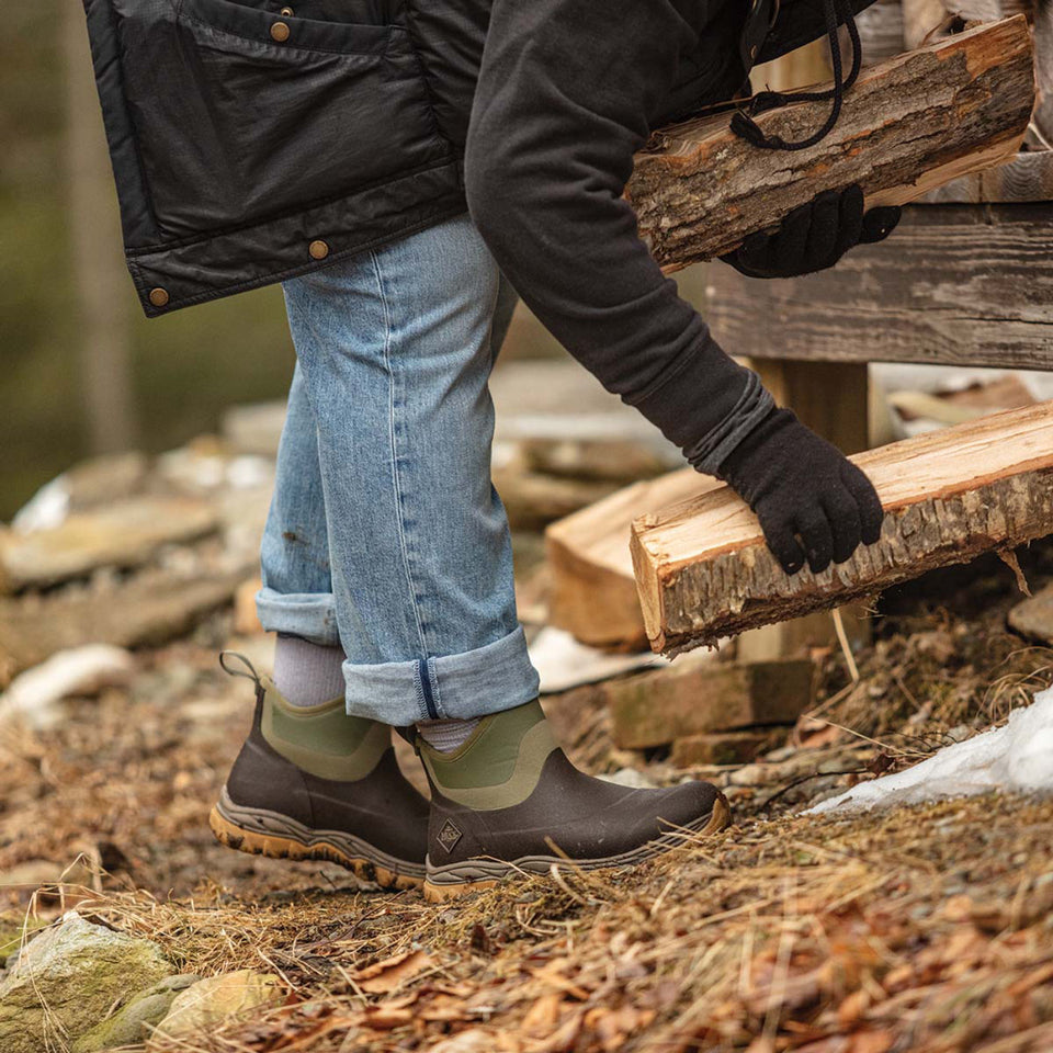 Person in black jacket, jeans, and brown boots carrying wooden logs outdoors.