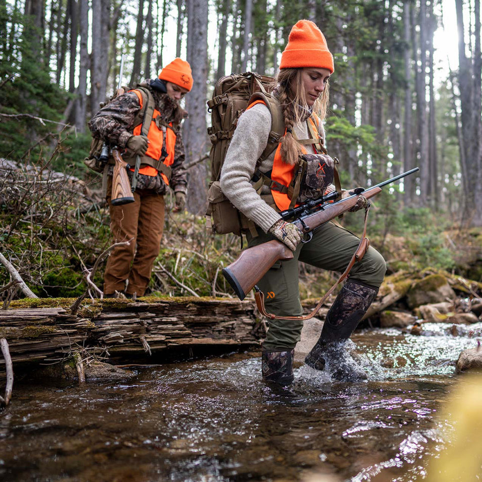 Two hunters crossing a stream in a forested area.