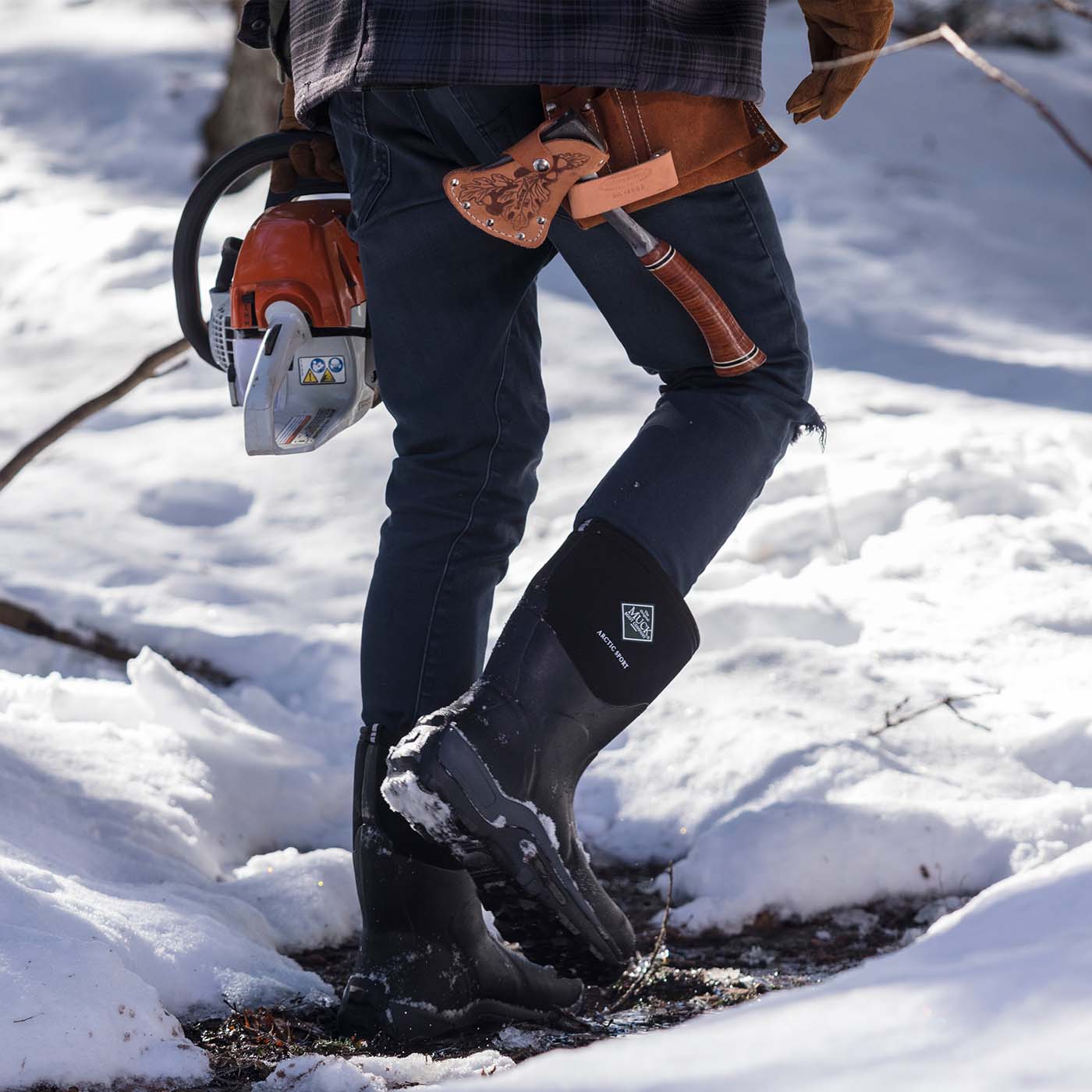 Person wearing black rubber boots with a chainsaw in a snowy environment
