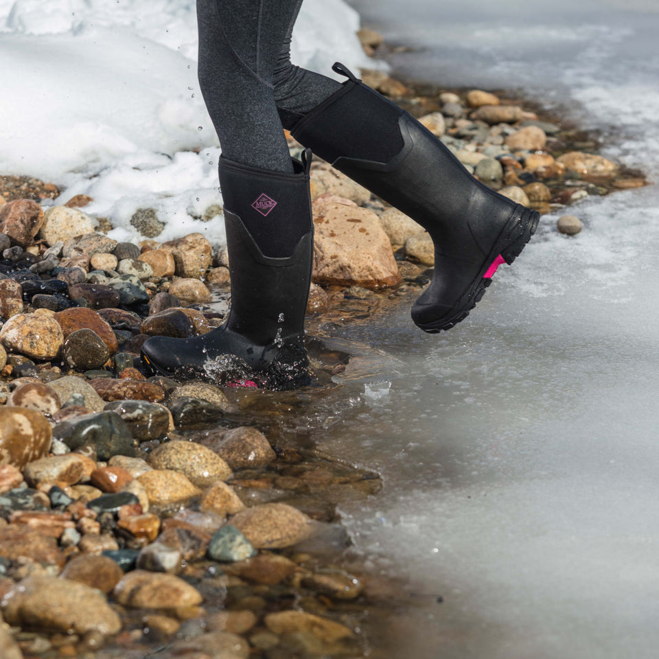 Person wearing black rubber insulated boots walking on a rocky shoreline with ice.