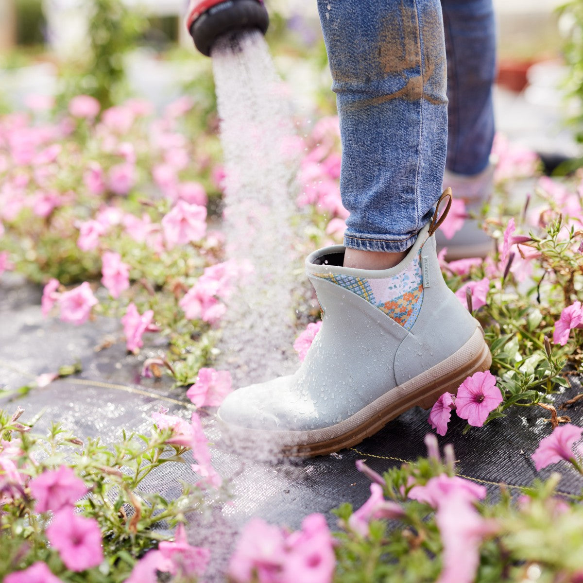 Women spraying her garden of pink flowers with water wearing Muck Boots.