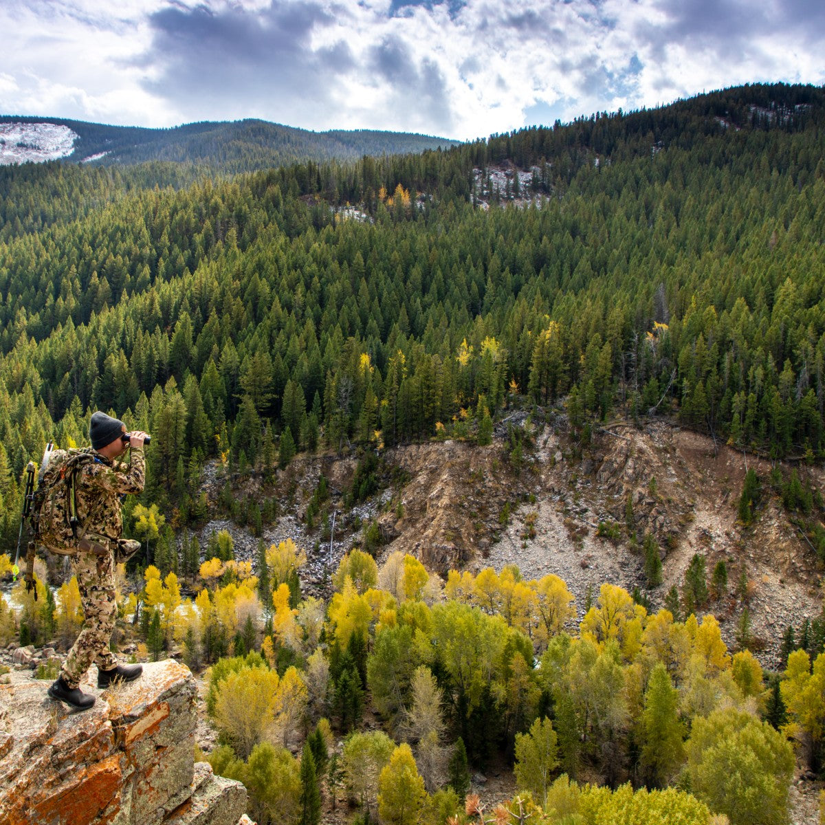 Hunter on rocky outcrop using his binoculars overlooking a forest.