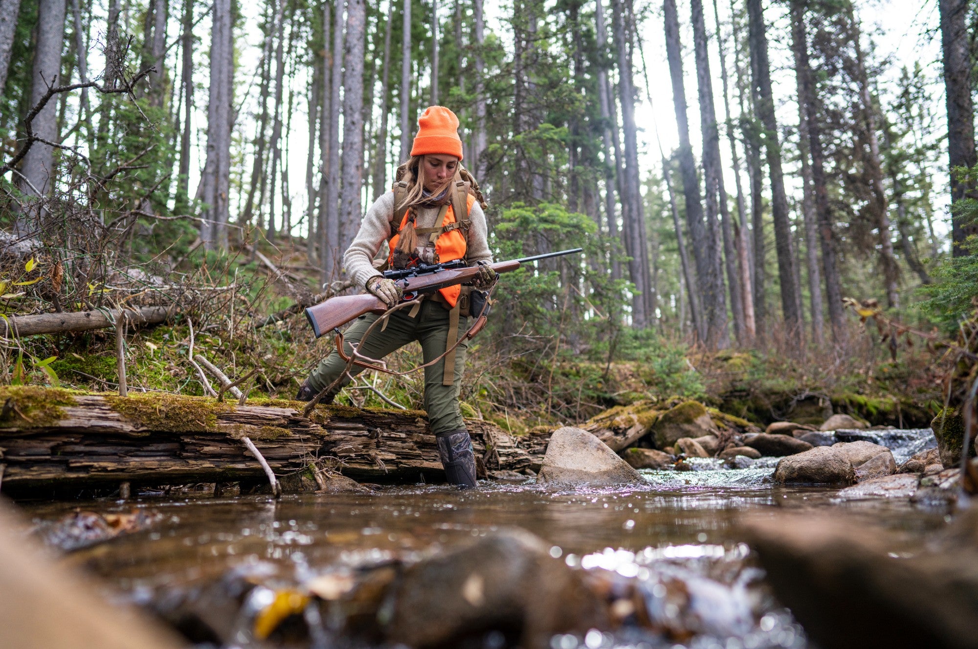 Women in hunting gear walking through stream in her Muck Boots.