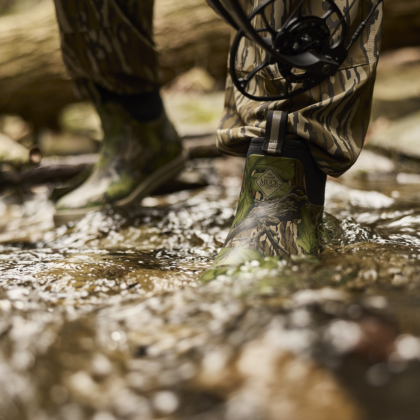 Person wearing camouflage boots walking through water, with a focus on the boot brand logo.