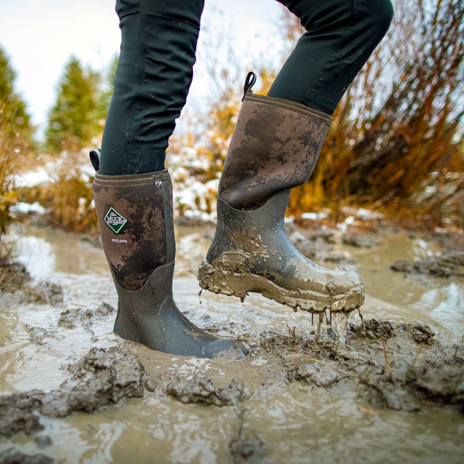Person wearing muddy rubber Wetland muck boots in a muddy outdoor setting with trees in the background.