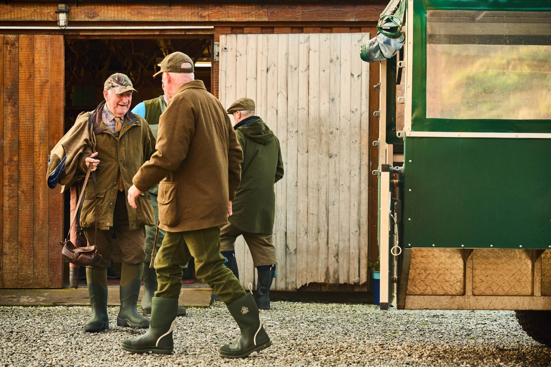 Three men in outdoor clothing walking towards a green vehicle near a wooden building wearing green chore muck boots