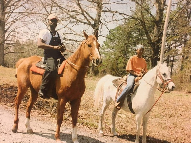 Two people riding horses on a dirt path with trees in the background