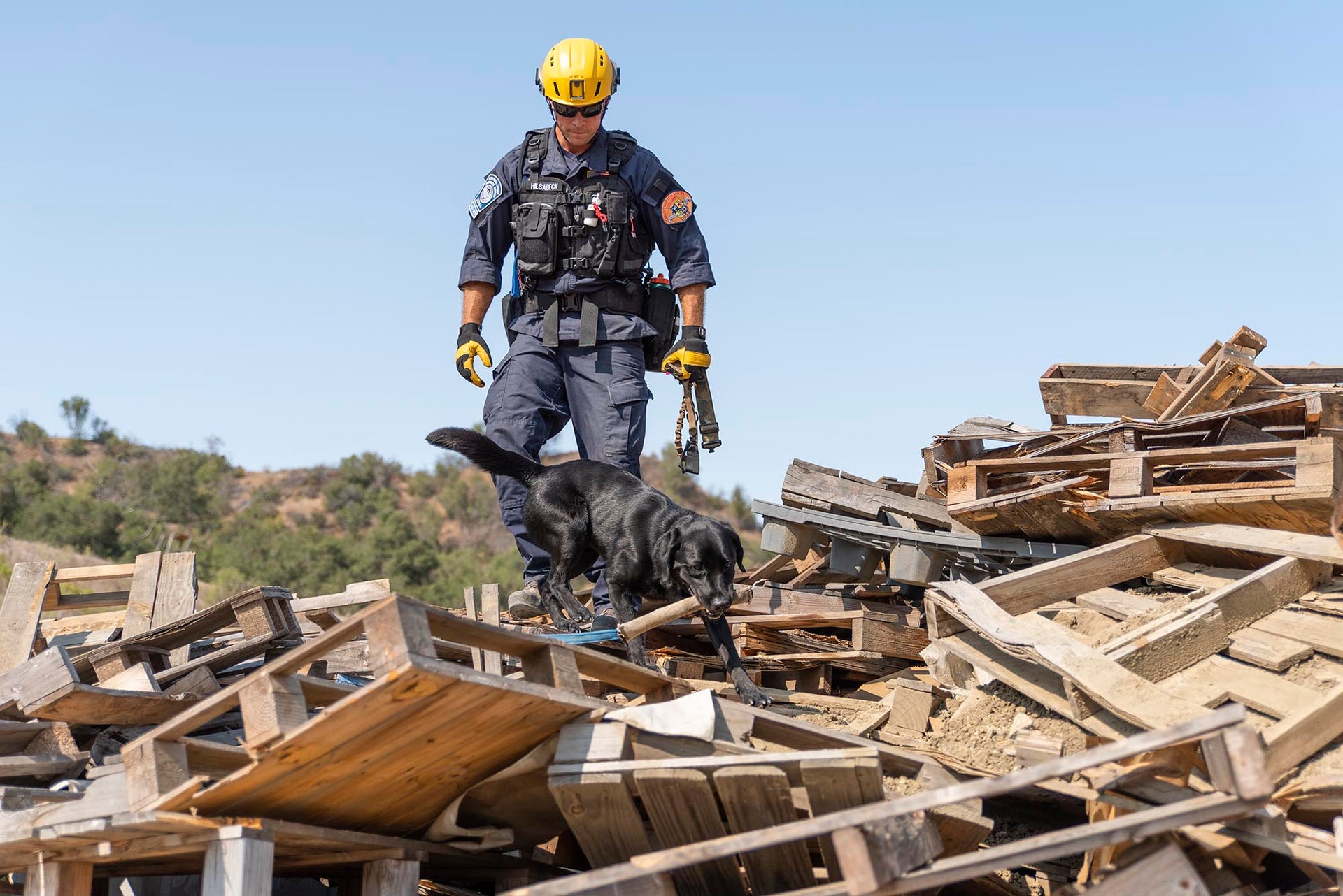 National Search Dog Foundation response team member in safety gear with a search dog during a rescue mission, on pile of wooden pallets
