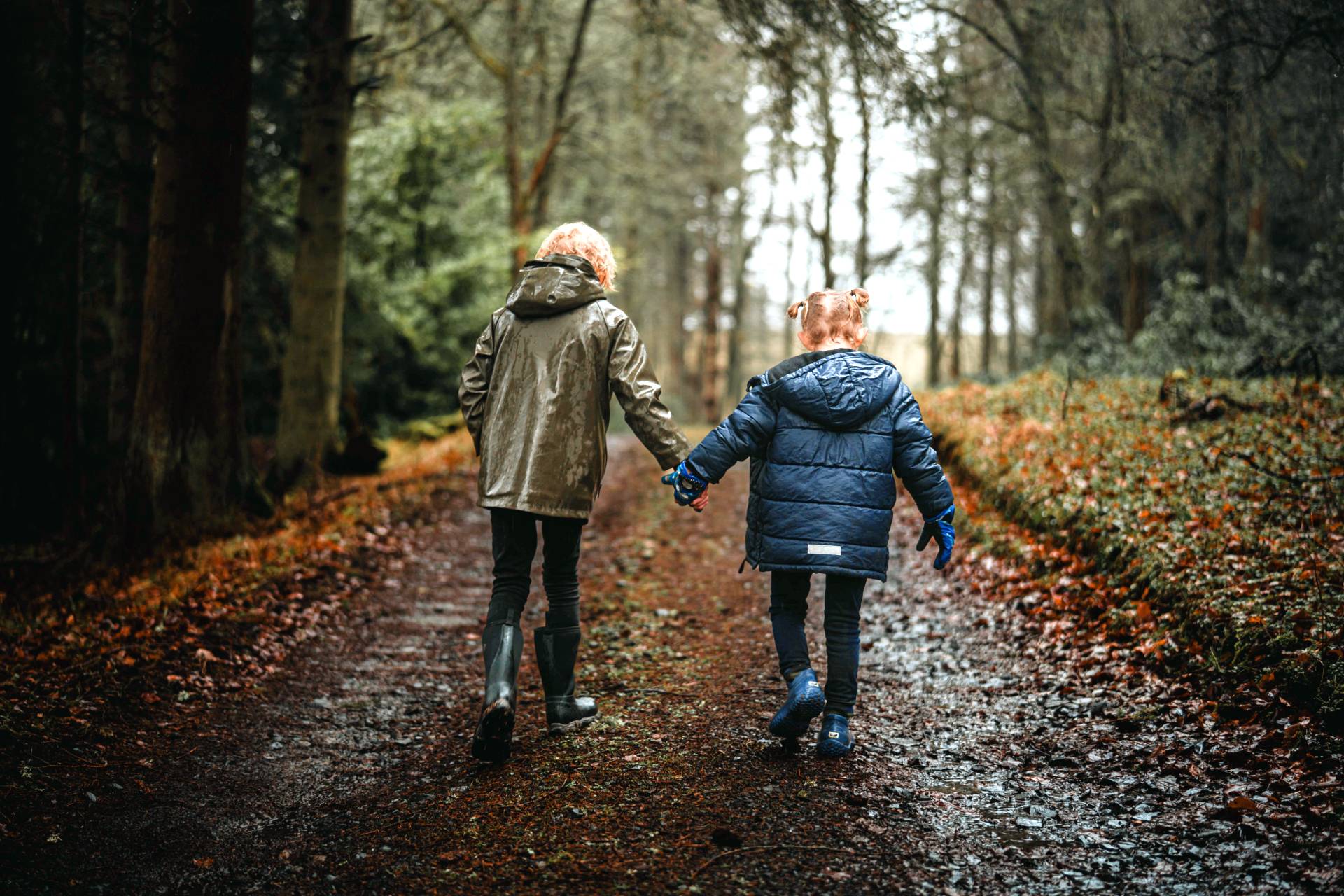Two children walking hand in hand along a muddy forest path, seen from behind. One child wears a beige hooded jacket and black rain boots, and the other wears a blue puffer jacket and blue rain boots.
