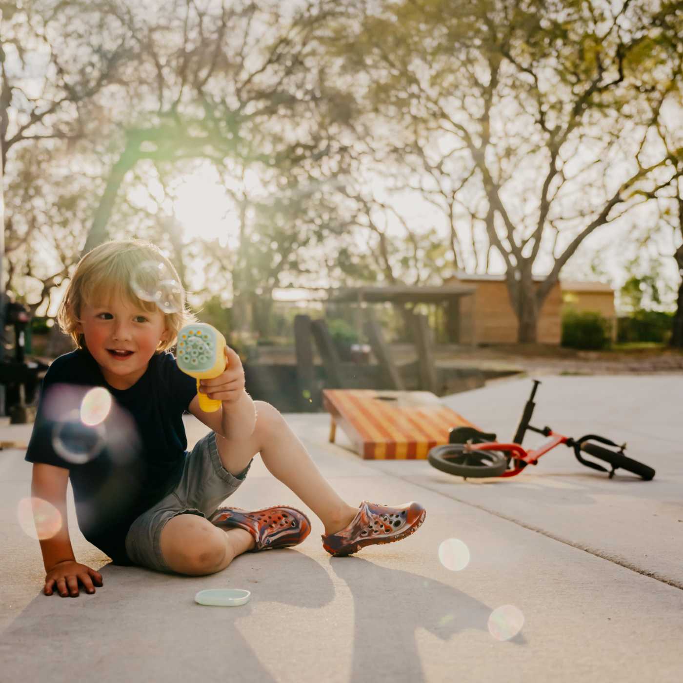 Child sitting on a driveway holding a bubble toy with a bike in the background. Wearing MMLCCUSS Muckster Lite Liberty Little Kids Clog