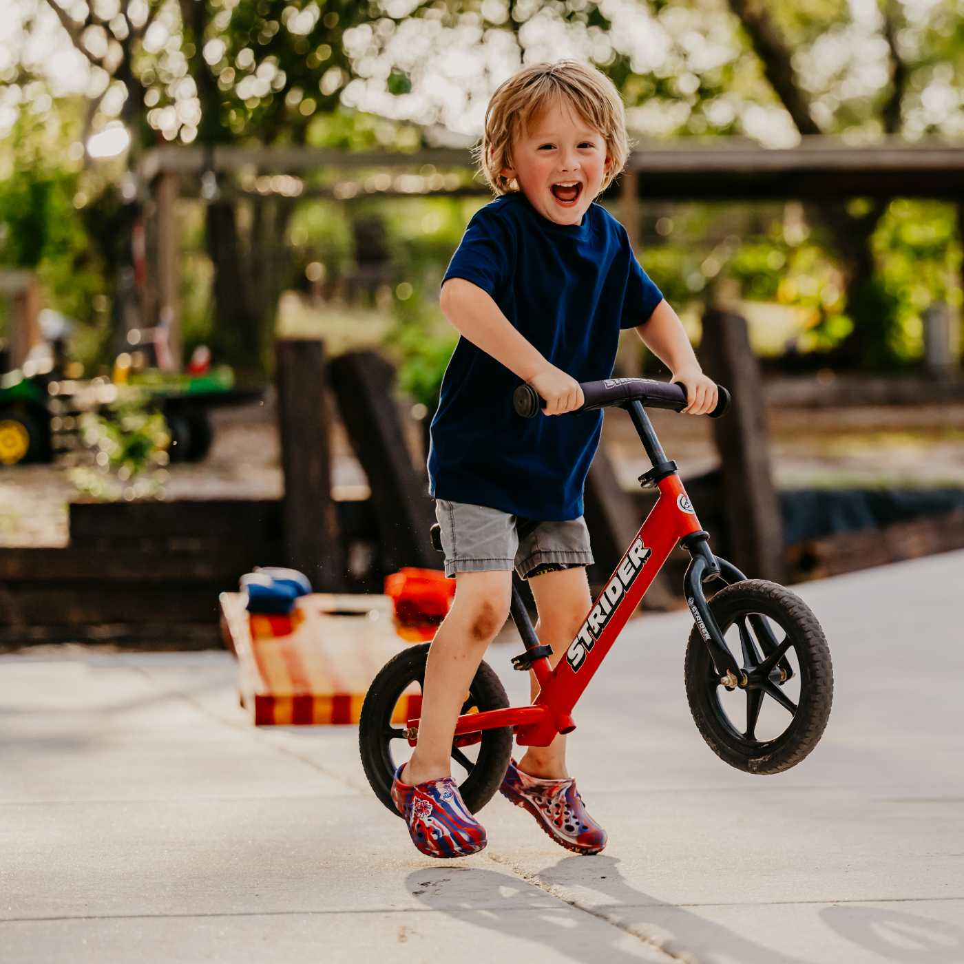 Child riding a red balance bike outdoors on a sunny day wearing MMLCCUSS Muckster Lite Liberty Kids Clog