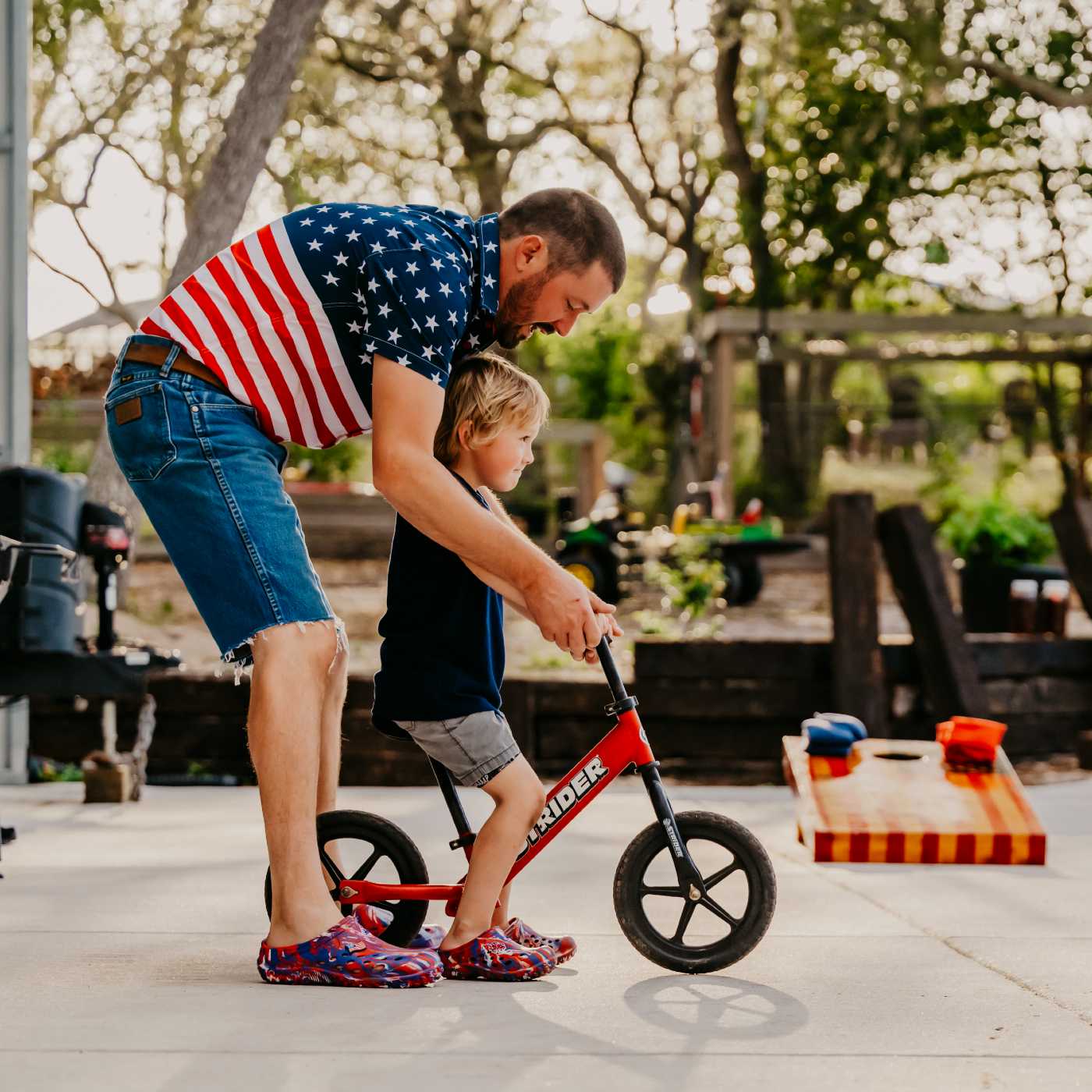 Man helping a child on a balance bike outdoors wearing Muckster Lite Liberty Clog MMLCMUSS