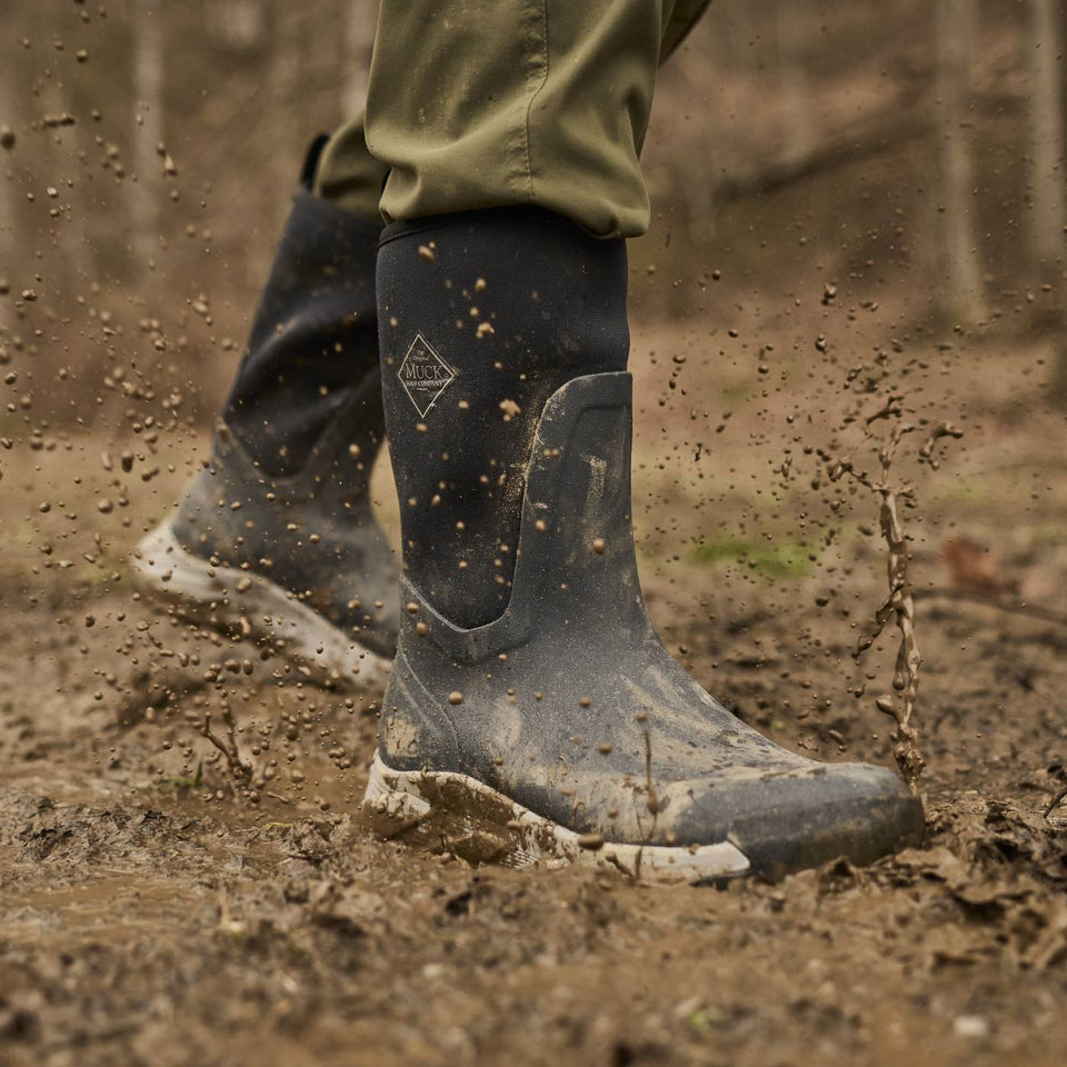 A close-up action shot of a person wearing dark gray rubber rain boots with mud splashing off them, standing in a muddy outdoor forest setting.