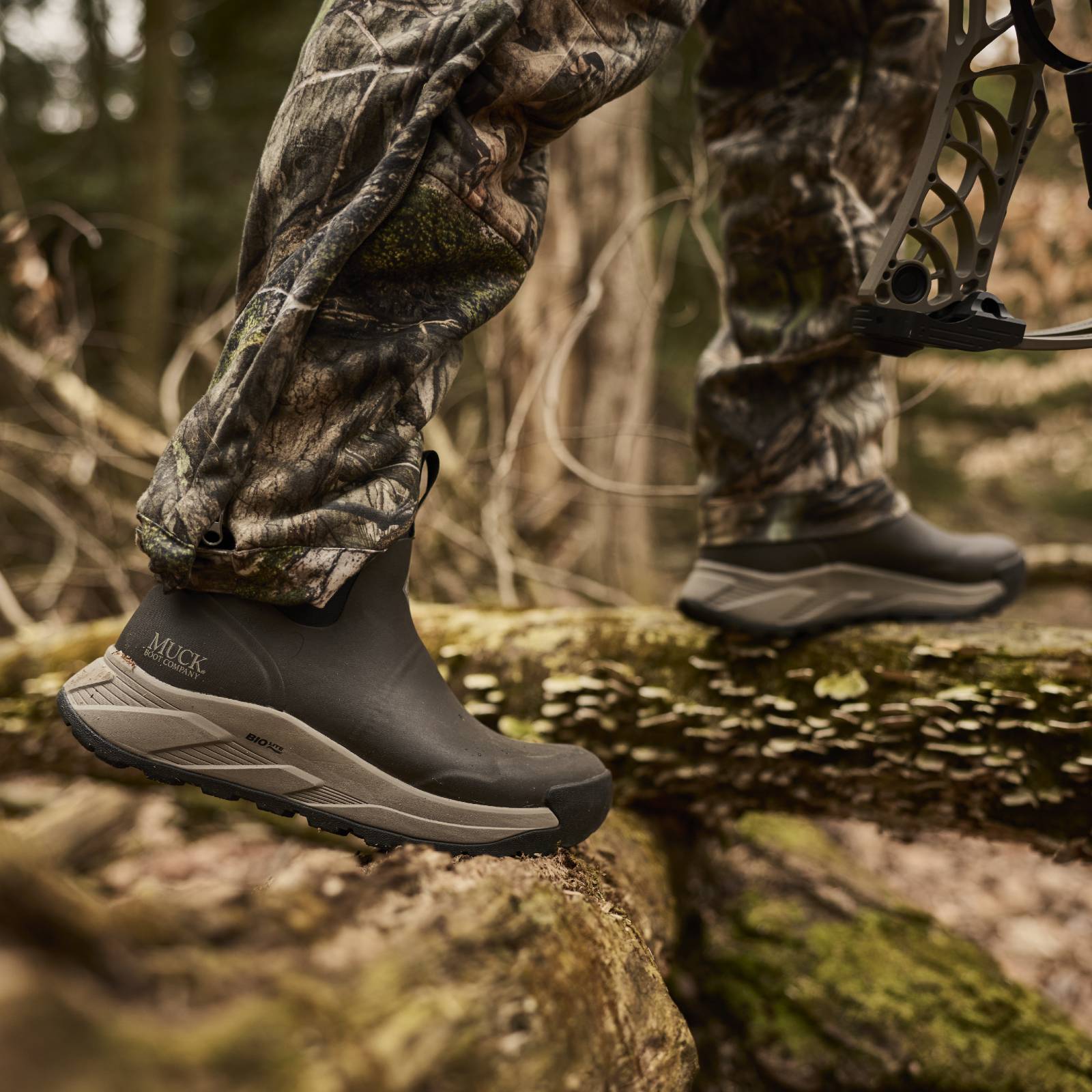A close-up outdoor shot of a person wearing camouflage pants and black Muck booties while stepping across a mossy log in a forest. A bow is partially visible on the right.