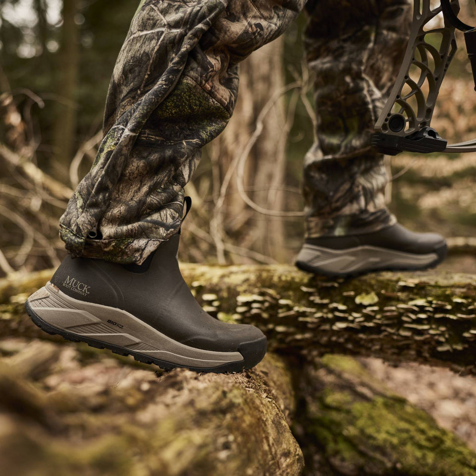 A close-up outdoor shot of a person wearing camouflage pants and black Muck booties while stepping across a mossy log in a forest. A bow is partially visible on the right.