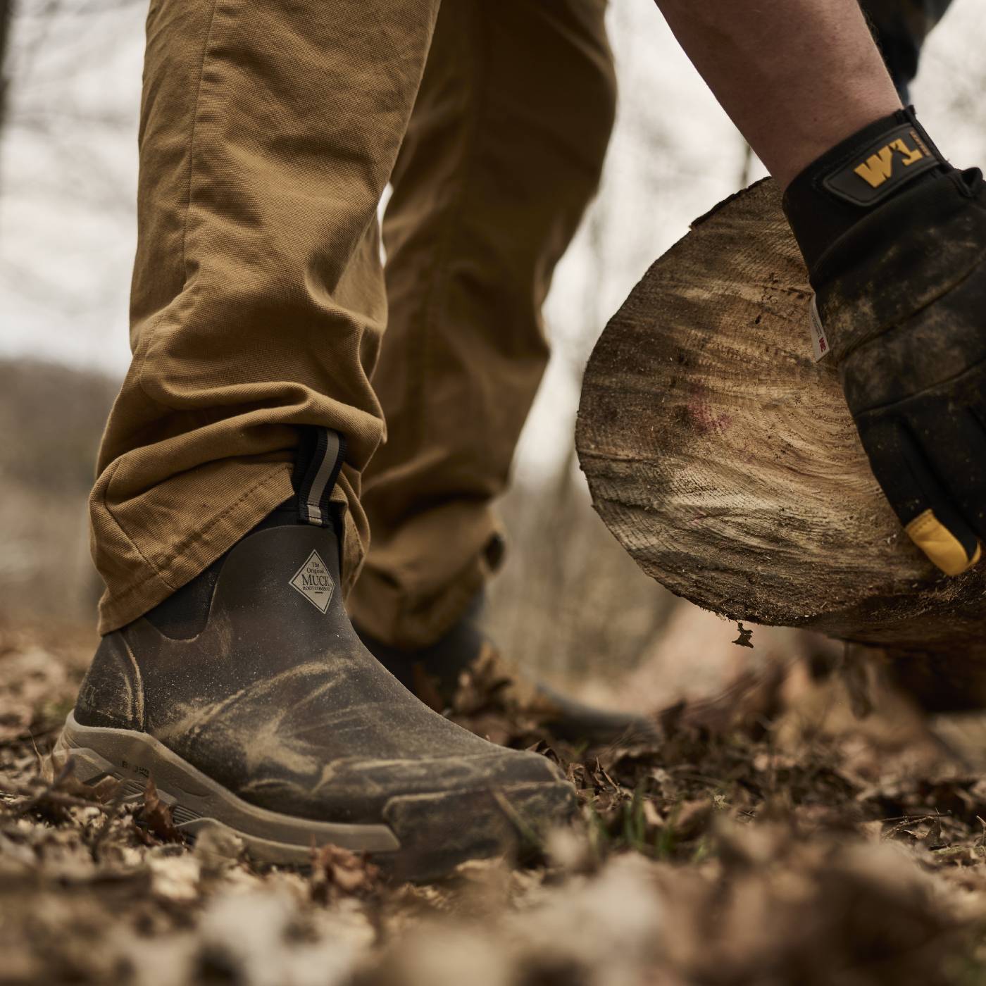 A close-up outdoor shot of a person wearing rugged waterproof boots and tan pants, standing on leaf-covered ground near a log while wearing a black work glove with yellow branding.