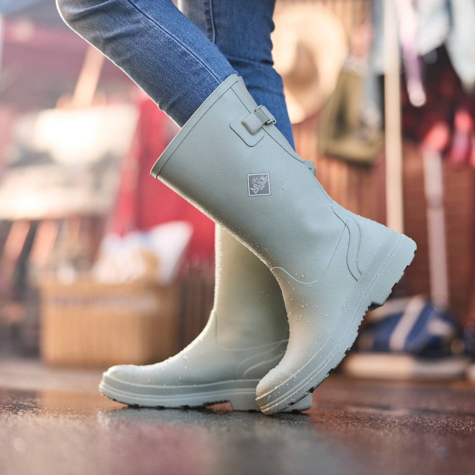 Tall green waterproof rubber boots worn with blue jeans on a blurred indoor background