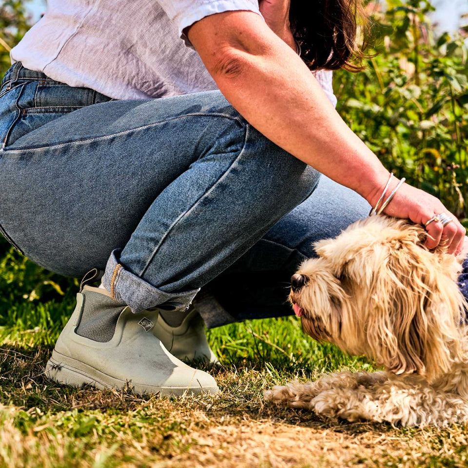 Person in jeans and light green waterproof rubber boots crouching on grass with a small dog.