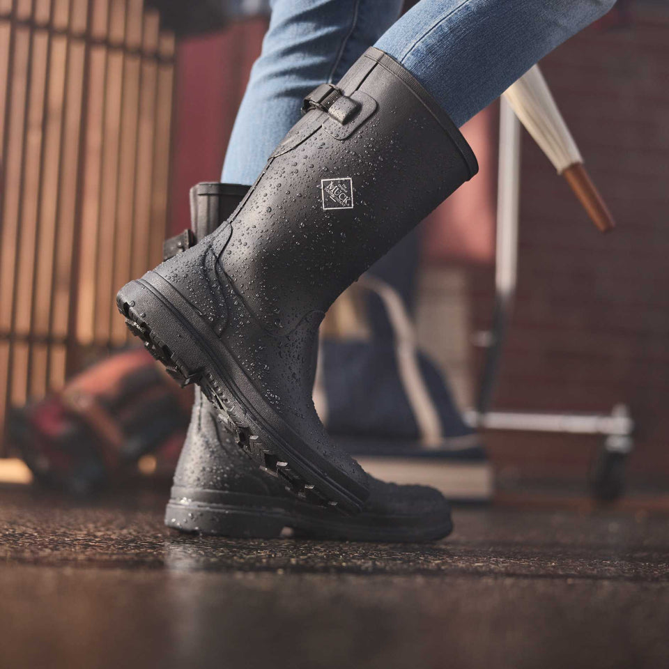 Waterproof black rain boots with a Muck Boot Co logo worn by a person, at a farmer's market on a rainy day