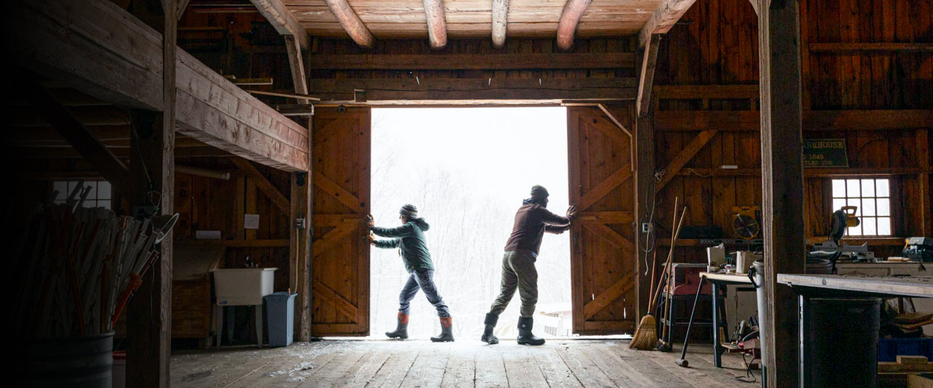 Two people entering a large wooden barn with tools and equipment inside.