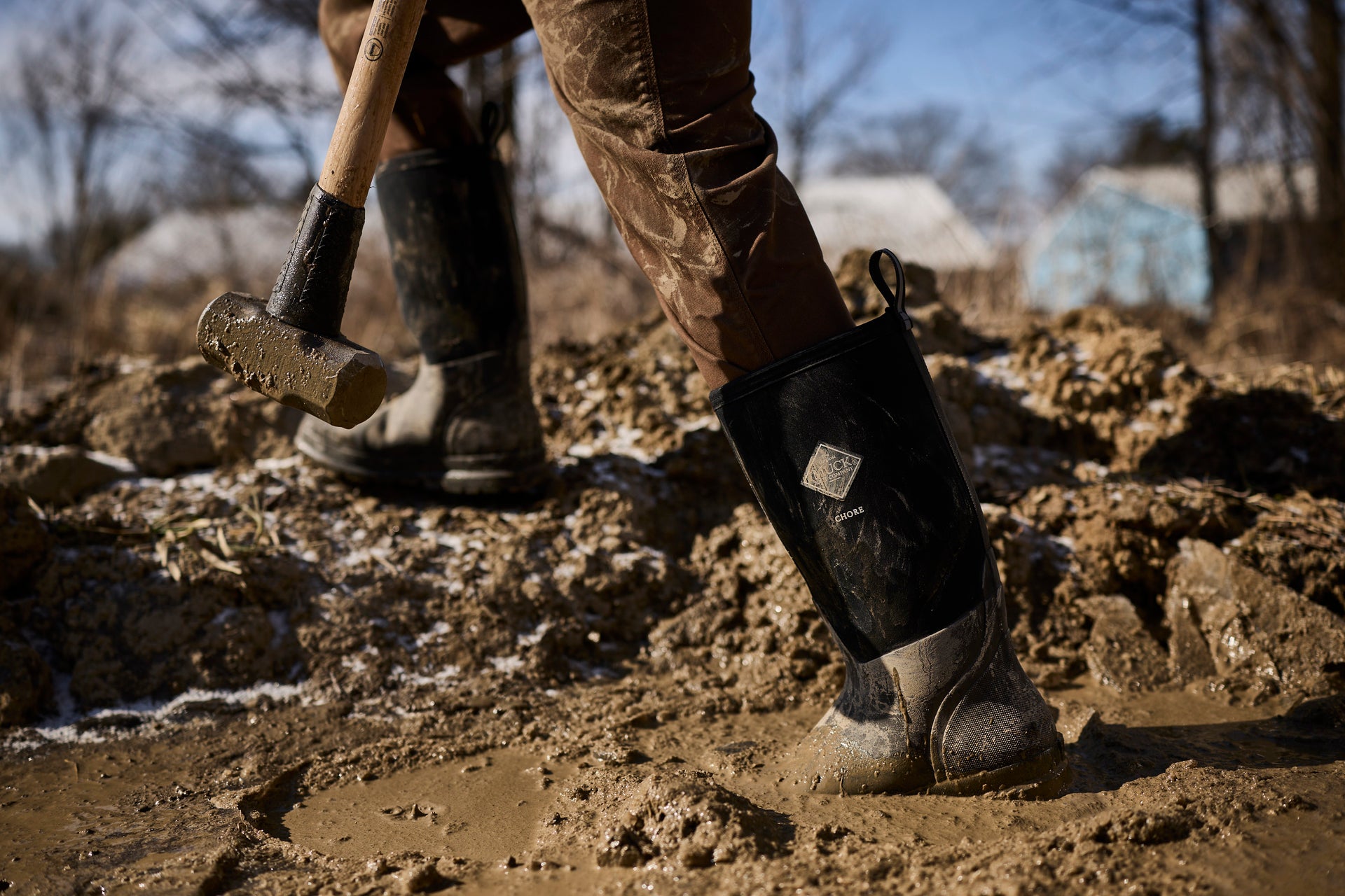 Person wearing black muck boots and brown pants walking through mud with a tool in a winter jobsite setting.