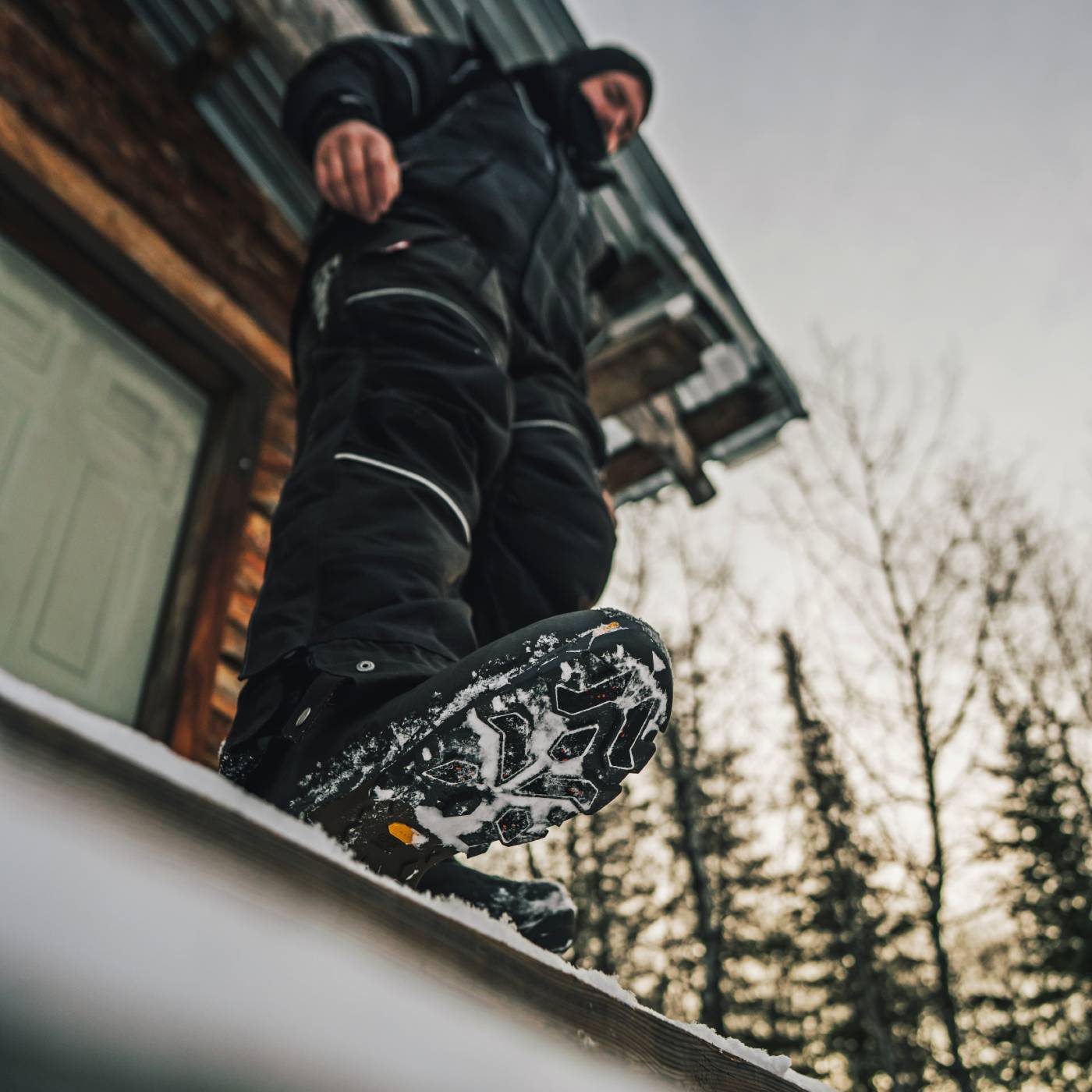 A person in black winter clothing steps onto a snowy roof in front of a wooden building, with bare trees and an overcast sky in the background. The product appears to be snowboard boots or winter footwear shown in use outdoors.