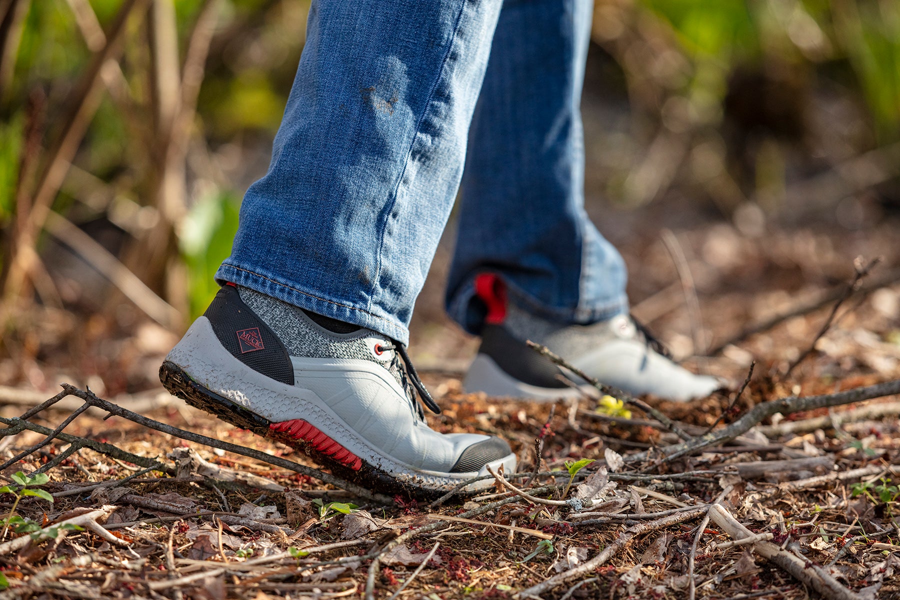 Person wearing gray waterproof shoes with red accents and blue jeans walking on a natural path.