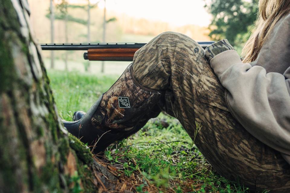 Person in camouflage muck boots with a rifle, sitting on the ground in a forest setting turkey hunting