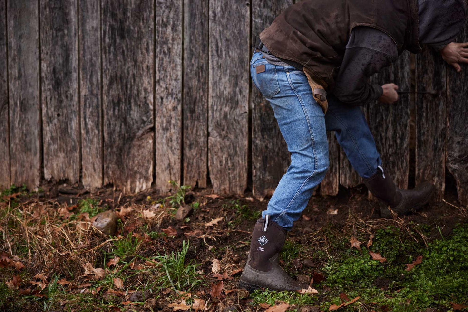 Person wearing blue jeans and brown muck boots working on a wooden fence.