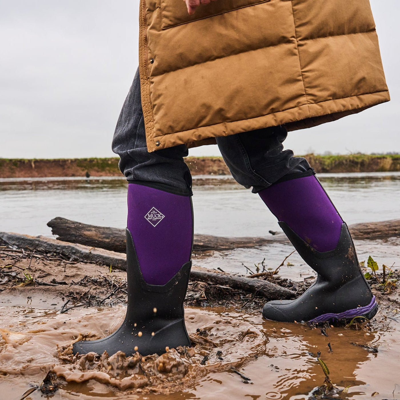 Person wearing purple boots and a brown jacket standing in a muddy area with water and logs.